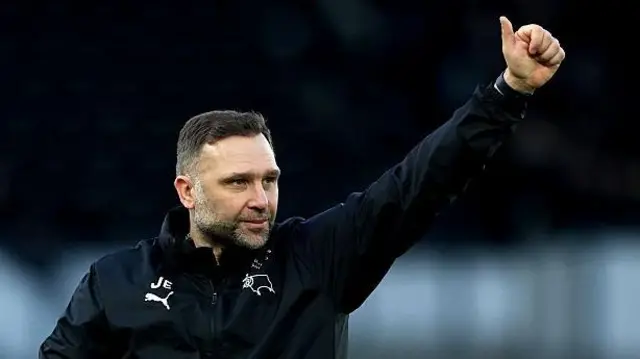 John Eustace, wearing a Derby tacksuit top, gives a thumbs up with his left hand to Derby supporters 