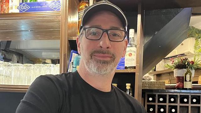A man with short grey facial hair, black glasses, a black cap and black t-shirt. He is smiling at the camera while stood behind a car in a restaurant
