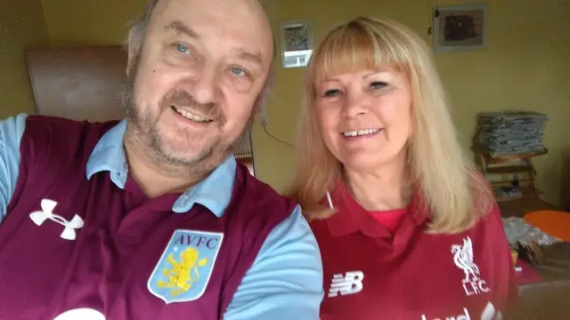 A man, who is dressed in an Aston Villa shirt, poses for a photo with his wife, who is dressed in a Liverpool shirt