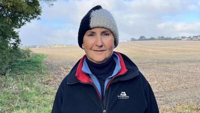 A woman in a navy fleece jacket and a black and white beanie. She is standing in front of an empty field.