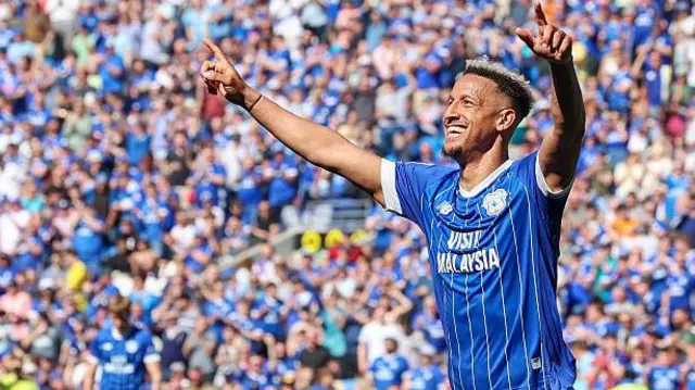 Callum Robinson in the blue and white of Cardiff City with arms raised aloft celebrating scoring a goal with other players and a packed crowd behind him