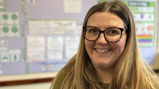 Angharad Pari-Williams smiles at the camera. She has rounded glasses and long blonde hair reaching past her shoulders. She is wearing a green top, while in the background a classroom display can be seen out of focus. 