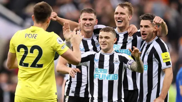 Newcastle outfield players Emil Krafth, Dan Burn, Fabian Schar and Kieran Trippier, wearing the black-and-white home shirt, stand smiling with their arms around each other as Nick Pope, with the back of his yellow top showing, walks towards them preparing to join in the celebrations.