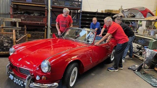 Four men leaning on the back of a shiny red convertible car with silver trim in a garage with rows of shelves with equipment behind them