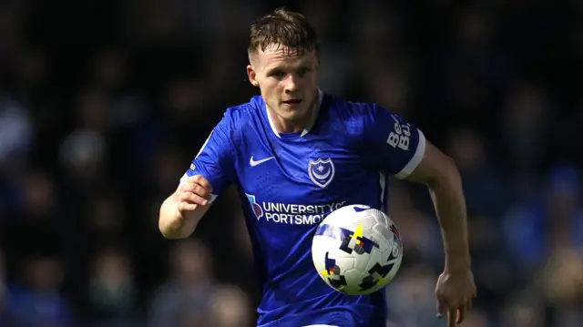 Terry Devlin, wearing Portsmouth's home blue shirt with a Nike tick and the club badge on the chest, looks towards the ball, which is in mid-air in front of him, during the home game against Ipswich. The Fratton Park crowd can be seen blurred out of focus behind him.