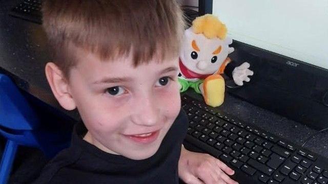 Leighton sitting at a computer is smiling at the camera with his puppet
