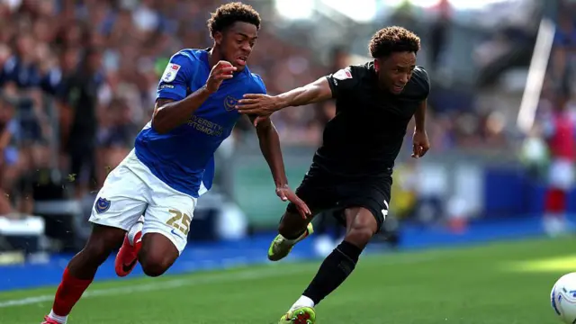 Kealey Adamson aggressively fends off Portsmouth defender with his right hand as he focuses on the white ball in front of him, wearing QPR all black away kit
