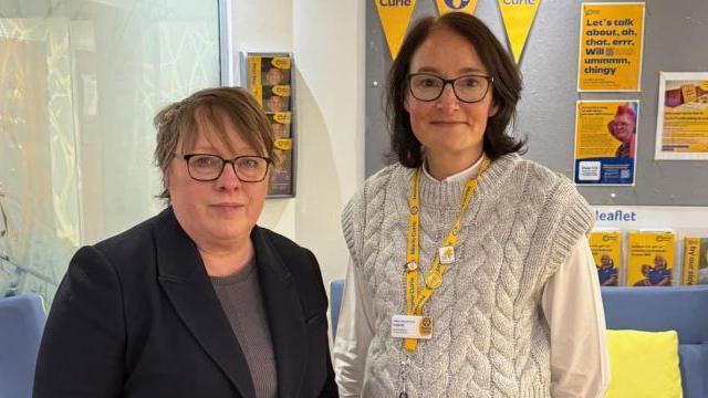 Maria Eagle has short hair, and is wearing dark rimmed glasses and a dark suit jacket and grey top. She is standing with another woman who has mid-length dark hair and is wearing dark-rimmed glasses and a cream coloured kitted top over a cream coloured shirt and a yellow lanyard. They are standing inside a building with Marie Curie leaflets behind them.