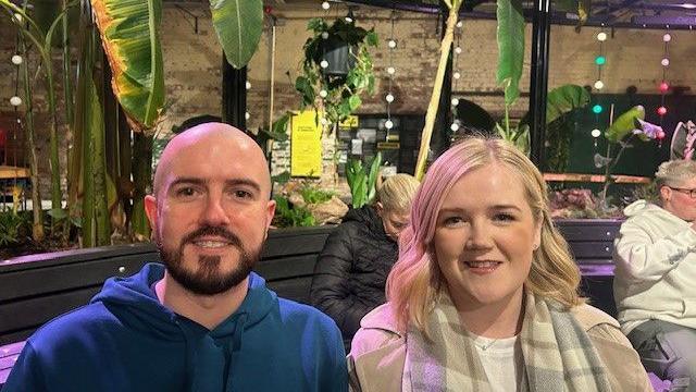 A man and woman are sitting down and smiling. There are plants and lights in the background. 