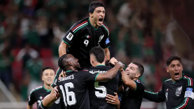 Raul Jimenez, wearing Mexico's black away shirt with red white and green Adidas stripes along the shoulders, celebrates their goal against South Korea at the World Cup with his team-mates by climbing on top of them. Edson Alvarez can be seen at the bottom right of the group of players with his mouth open celebrating.