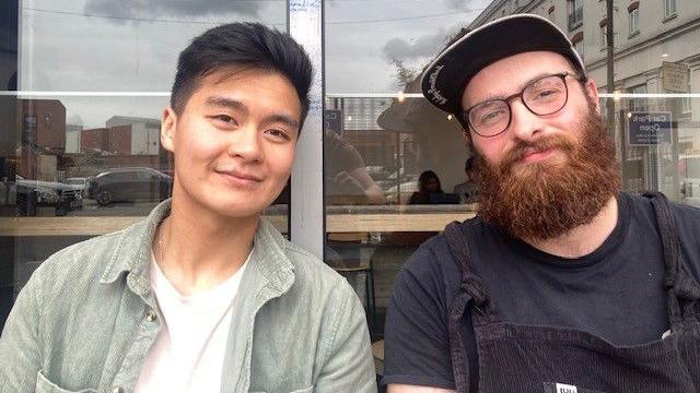 Two people smile at the camera. On the left is a man in a white top and a light green jacket. On the right is a man with a beard and glasses, wearing a navy t-shirt and black baseball cap.