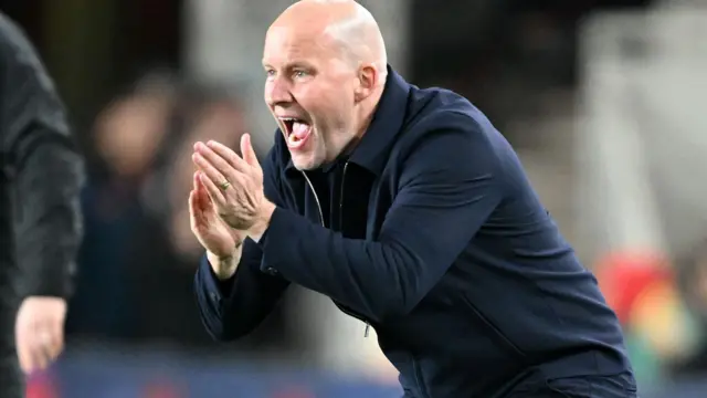 Henrik Pedersen crouches on touchline, hands clapping encouragement as he shouts instructions, wearing all navy blue trousers and zipped warm jacket.