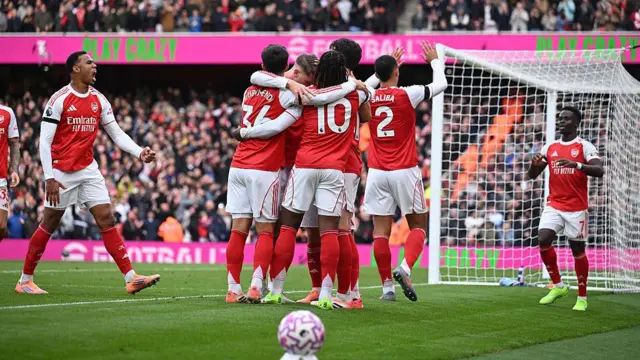 Several Arsenal players celebrate a goal in a huddle. Gabriel is to the side with his fist clenched in celebration towards the crowd and Bukayo Saka is coming to join the huddle. The crowd at Emirates Stadium is a blur in the background.