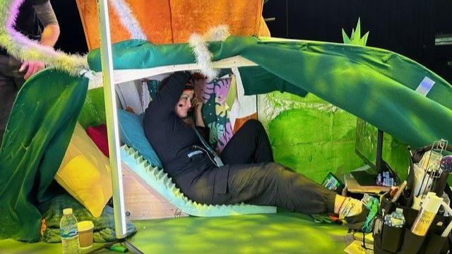 A puppeteer is showing sitting underneath a TV set as she operates her puppet on a grassy mound