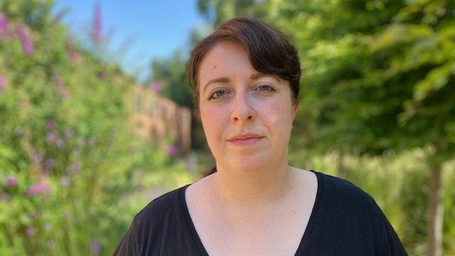 Shauna Vincent has short brown hair and is standing in a garden wearing a v-neck black top looking into the camera