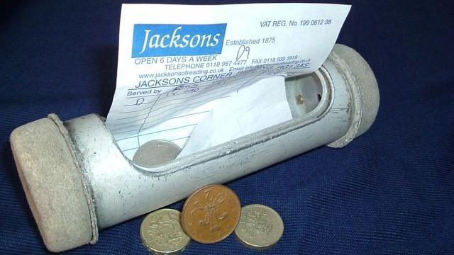 A glass canister which is open and has a piece of paper which says "Jacksons" and the details about the purchase. There are two pound coins, a two pence piece and a ten pence piece.