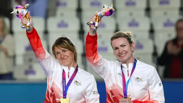 England pair Amy Pharaoh and Sian Honnor smiling with their gold medals around their neck, holding their mascots from the Glasgow 2026 Commonwealth Games in the air