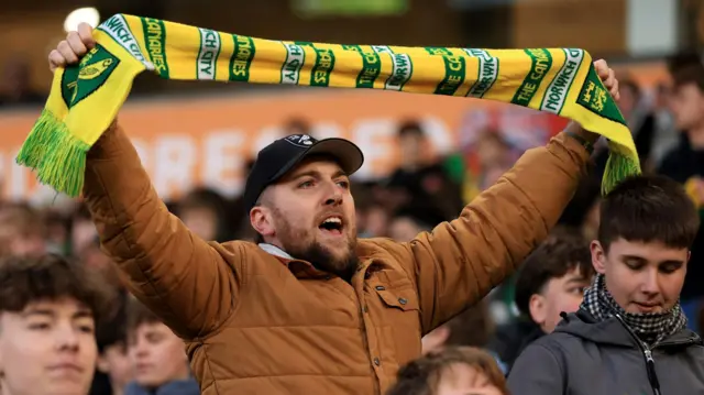 Supporter wearing a brown jacket and black baseball cap holds a green and yellow Norwich City scarf above his head during their FA Cup tie against West Bromwich Albion
