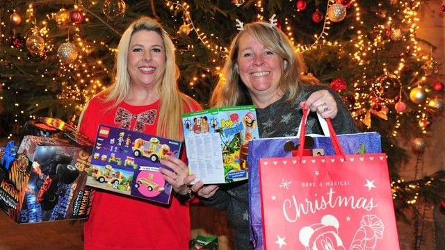 Natalie, a woman with gold hair, wearing a red T-shirt holding toys in her hand, smiling for camera and Sharron, a women with dark golden hair also holding Christmas gift bags. Both of them are standing in front of a Christmas tree.