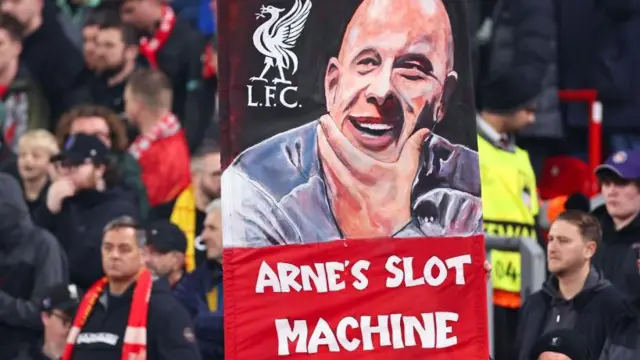 Liverpool fans hold up a banner reading "Arne's Slot machine" with a depiction of the Dutchman smiling above