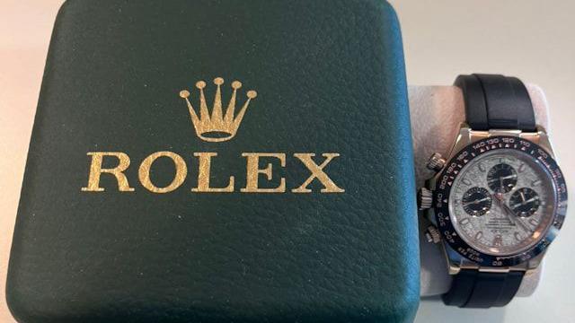 A green Rolex watch box which says 'Rolex' on the front in gold lettering, to the right, a black strap Rolex watch