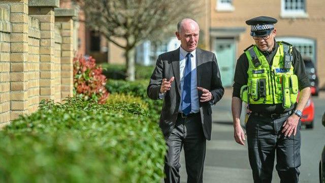 Darryl Preston, wearing a black suit and blue tie, is walking next to a male police officer wearing a yellow hi-vis jacket. They are in conversation, on a residential street in Peterborough.