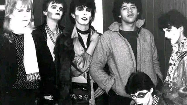An old black-and-white photo of five punk rockers in black-and-white face makeup. Another man stands with his hands in his jacket pocket.