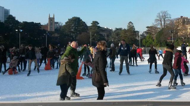Scores of people wrapped up in coats and hats, skating on an outdoor ice rink on a sunny day. The scene is set against a backdrop of trees with a church tower poking over the tree line in the distance.
