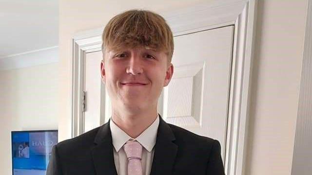 Alfie Gray smiles at the camera whilst wearing a black suit, white shirt and pink tie