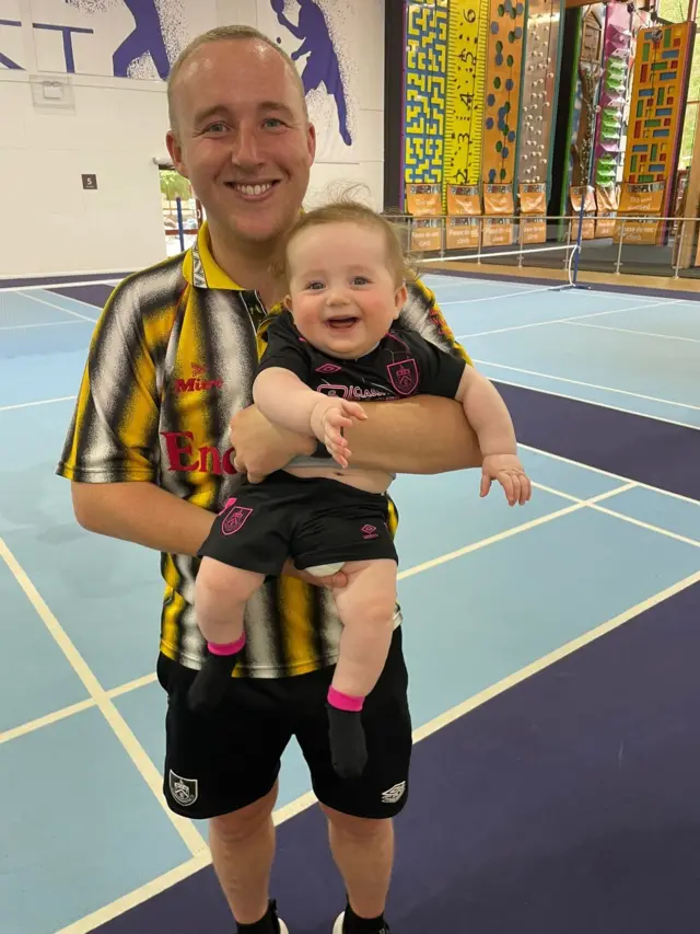 A man in a yellow and black Burnley kit holds a baby in a Burnley strip