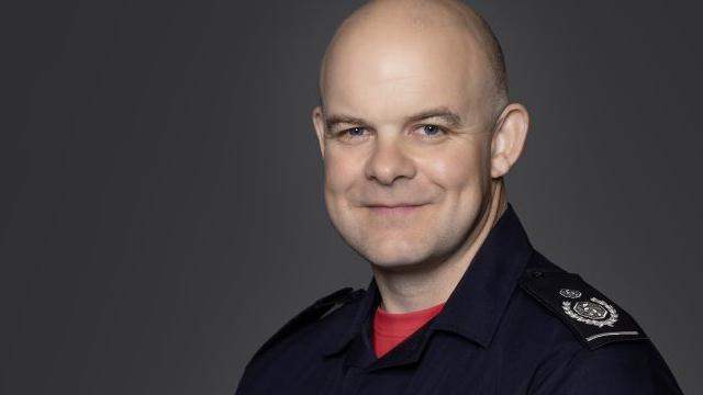 Formal work photo of Jonathan Smith, a bald man wearing a red T-shirt under a navy fire brigade boiler suit. There is a plain background and he is smiling.