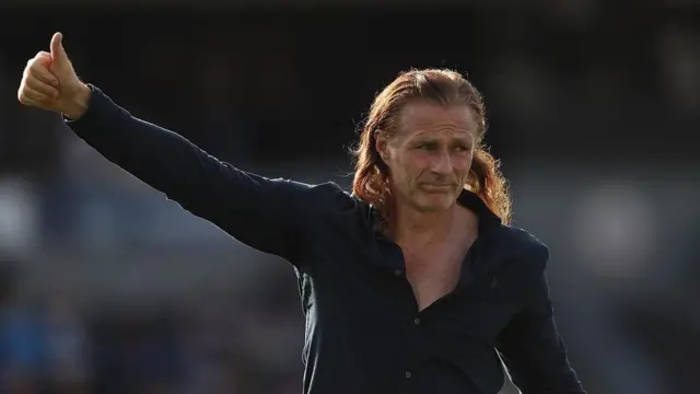 Gillingham manager Gareth Ainsworth walking across pitch with right thumb held up to the fans, wearing a blue long-sleeve shirt unbuttoned to his chest