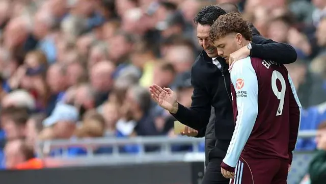 Unai Emery with his arm around Harvey Elliott's shoulder as he gives the player instructions on the touchline during a match. The crowd is out of focus in the background.
