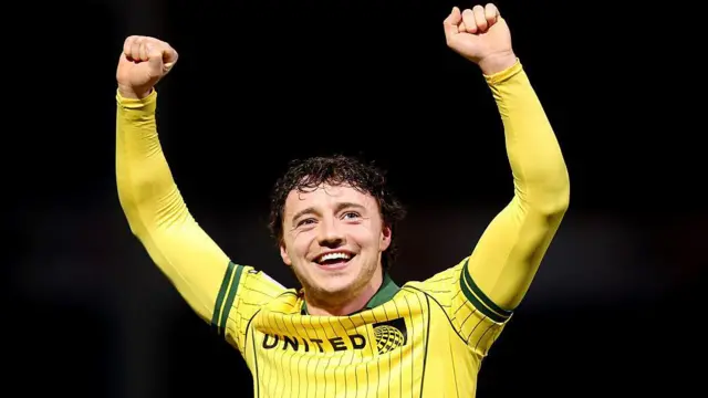 Ollie Rathbone smiles with his arms above his head wearing a yellow Wrexham away shirt