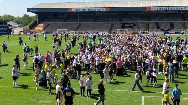 Hundreds of fans on a football pitch. They are on green grass and there is a stand behind them.