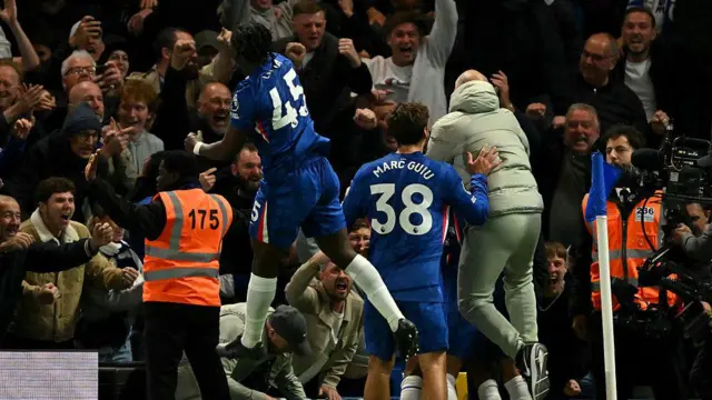 Enzo Maresca celebrating with a huddle of Chelsea players after the winner against Liverpool, as Romeo Lavia leaps into the air and punches the air towards the crowd of supporters, who are all smiling and celebrating in the background