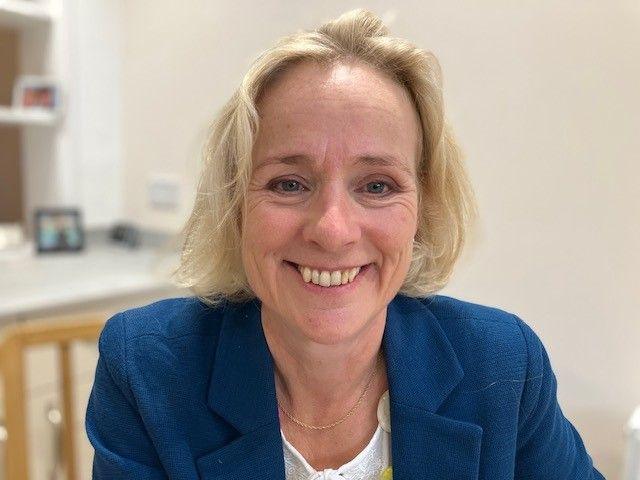 Former MP Vicky Ford smiles at the camera. She has shoulder-length blonde hair and is wearing a blue jacket and a white top. The background is out of focus, but could be an office.