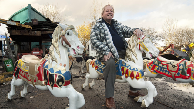 Jimmy Stringfellow in a thick checked shirt and grey trousers, sits astride a carousel horse with several more on either side of him, within his yard.