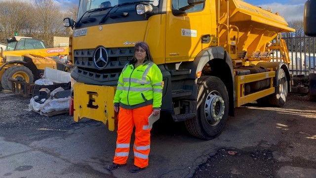 A woman who is a gritter driver dressed in orange and yellow hi-vis stands next to a gritter 