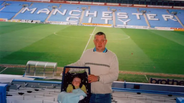 A picture of a man holding a baby in a car seat at Maine Road