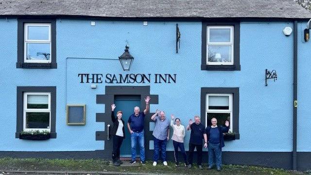 Tenant George Campbell waves at the camera as he stands outside the pub with Steve Rozario, vice-chairman of the Save Our Samson campaign, and four other members of the community. The pub is painted light blue with a darker trim around the windows and door. A sign and lamp hang above the door.