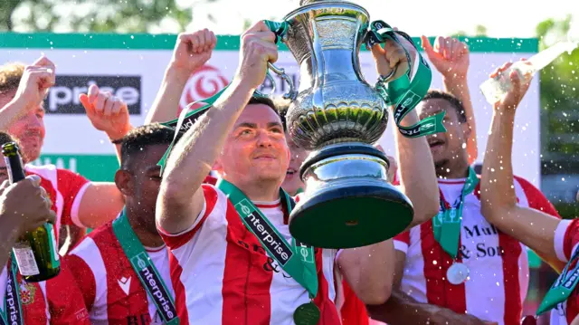 Hornchurch's Tom Wraight, in red and white striped kit, lifts the National League South play-off trophy, with his team-mates around him