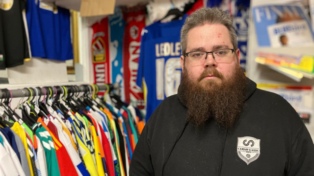 Daniel Rees standing beside a rack of multi-coloured shirts and in front of a wall displaying team shirts.