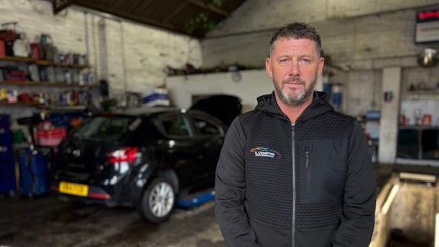 A man is standing in a motoring garage wearing a black jacket. There is a black car in the background.