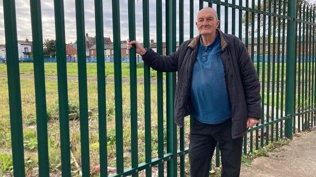 Billy Marrat, wearing a blue jumper under a dark blue coat, leans on a green metal fence