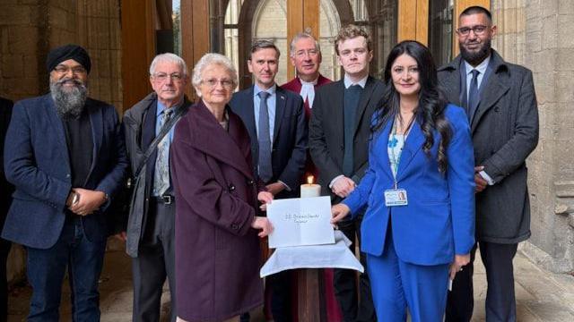 Members of the public including a man in a turban, a man in a coat and trousers, a Janet wearing a purple coat,  Andrew Pakes and Sam Carling- wearing suits , Tim Jones wearing a red ceremonial robe, Shabina wearing a blue suit and Zameer Ali wearing a grey coat- standing in a group for the pic.