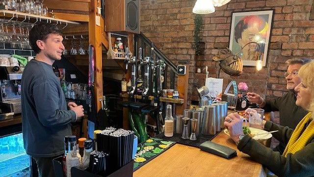 Bar owner James Ince pictured in a dark blue top, behind a bar speaking to two customers a male and female. 