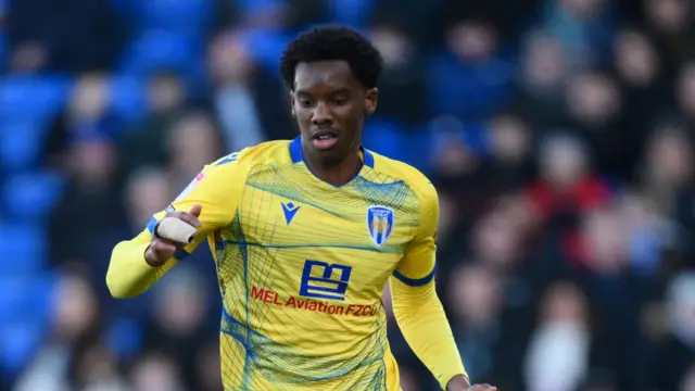 Harvey Araujo playing for Colchester United in their yellow kit, with two of his fingers taped together