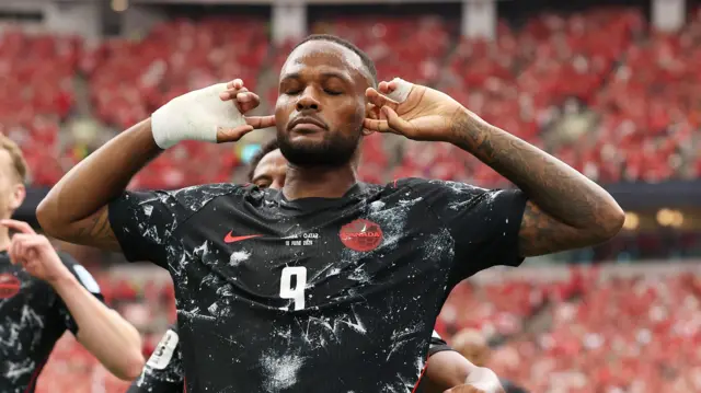A photo of Cyle Larin, wearing his black Canada kit, holding his fingers to his ears with his eyes closed in celebration of his goal against Qatar on 18 June