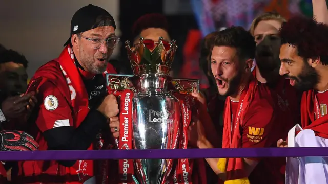 Jurgen Klopp, wearing a red Liverpool shirt over his dark top and a backwards cap, preparing to lift the Premier League trophy with Adam Lallana and Mohamed Salah